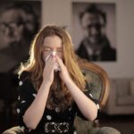 A young woman with closed eyes sneezing into a tissue, sitting indoors.