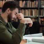 A tired man with glasses in a library, holding his head, symbolizing stress or fatigue.