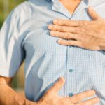 Close-up of a man in a blue shirt holding his chest and stomach outdoors, indicating discomfort.